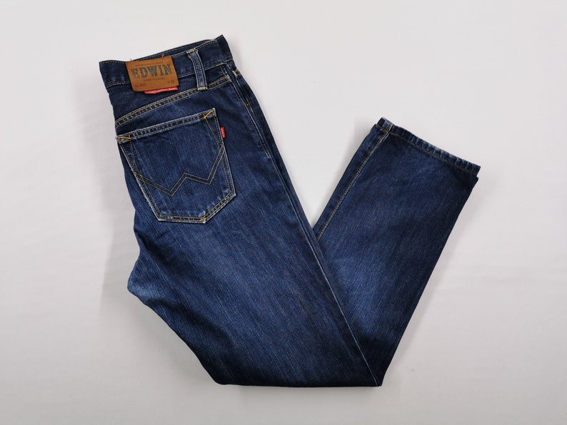 May include: Dark blue denim jeans with a classic five-pocket design. Features a brown leather patch with the brand name "EDWIN" on the back waistband. The back pocket has a unique zigzag stitch detail. The jeans are laid flat.