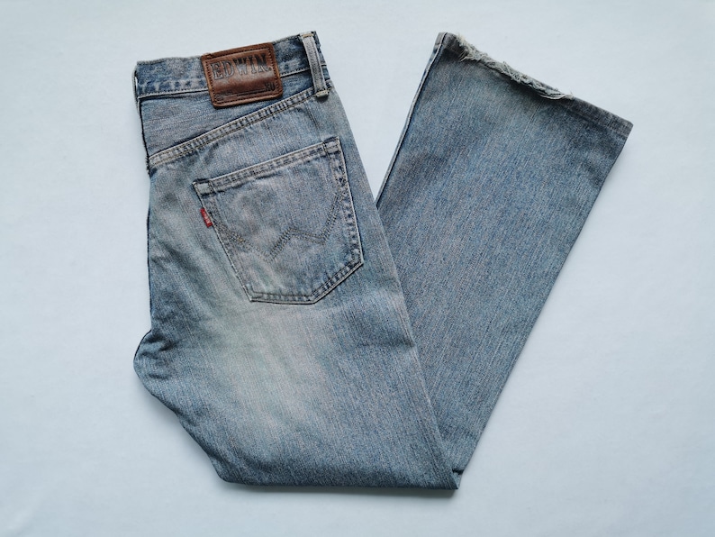 May include: Faded light blue denim jeans with a classic five-pocket design. Features a leather patch with the brand name "Edwin" and a red logo tag. The jeans have a straight leg and a worn appearance. The jeans are made of denim.