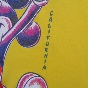 Op de afbeelding: Mosterdgeel T-shirt met een Mickey Mouse cartoonontwerp in paars, wit en rood. Het woord "CALIFORNIA" is verticaal in groen gedrukt. Het ontwerp staat aan de linkerkant van het shirt.