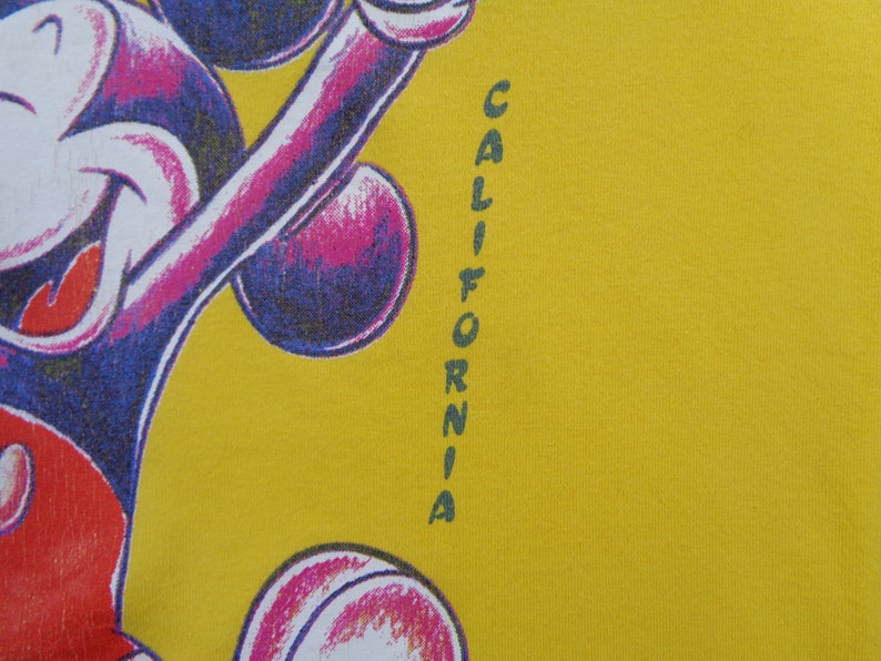 Op de afbeelding: Mosterdgeel T-shirt met een Mickey Mouse cartoonontwerp in paars, wit en rood. Het woord "CALIFORNIA" is verticaal in groen gedrukt. Het ontwerp staat aan de linkerkant van het shirt.