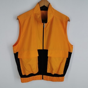 May include: A sleeveless orange and black vest with a full-length zipper. The vest has two front pockets and a black mesh panel on the sides. The vest is hanging on a wooden hanger.