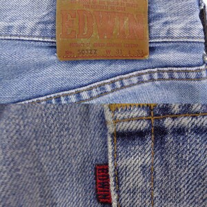 May include: Close-up of light blue denim jeans with a brown leather patch featuring the brand name "EDWIN" in red lettering. The jeans have a red tag with "EDWIN" stitched on the back pocket. The jeans are size W31 L33.