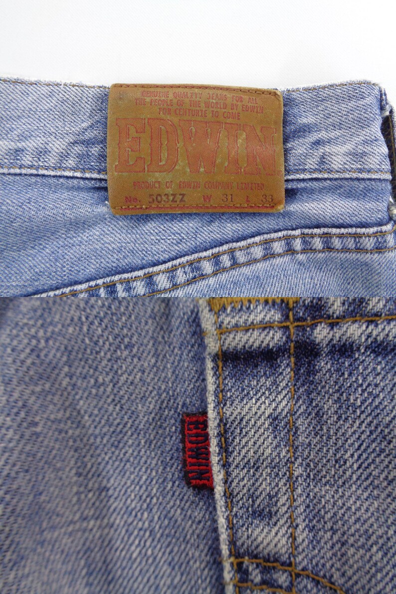 May include: Close-up of light blue denim jeans with a brown leather patch featuring the brand name "EDWIN" in red lettering. The jeans have a red tag with "EDWIN" stitched on the back pocket. The jeans are size W31 L33.