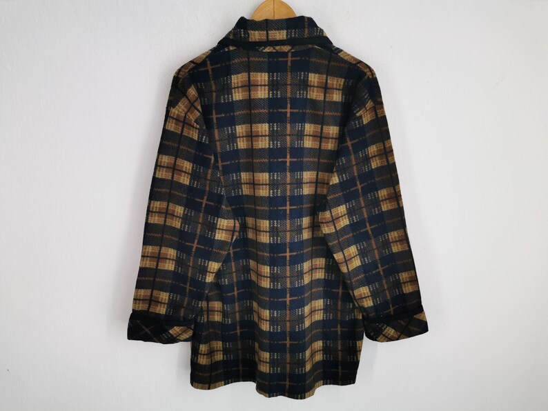 Puede incluir: Camisa de manga larga con botones y estampado de cuadros en tonos marr&oacute;n, azul marino y beige. La camisa tiene cuello y pu&ntilde;os abotonados. La parte trasera de la camisa es visible, colgada de una percha de madera.