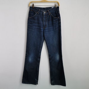 May include: Dark wash denim jeans hanging on a wooden hanger. The jeans feature a classic five-pocket design, a button and zip closure, and a slight bootcut leg opening. The denim shows some fading and wear.