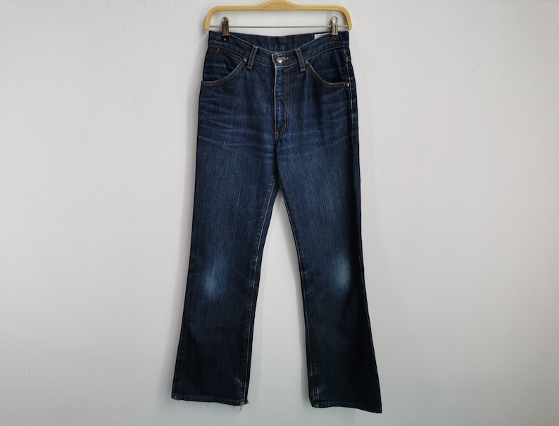May include: Dark wash denim jeans hanging on a wooden hanger. The jeans feature a classic five-pocket design, a button and zip closure, and a slight bootcut leg opening. The denim shows some fading and wear.