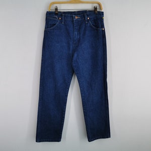 K&ouml;nnte beinhalten: Dunkelblaue Jeans aus Denim, die an einem Holzb&uuml;gel h&auml;ngen. Die Jeans haben ein klassisches F&uuml;nf-Taschen-Design mit geradem Bein. Der Denim scheint ein mittleres Gewicht zu haben, geeignet f&uuml;r den t&auml;glichen Gebrauch. Die Jeans werden vor einem schlichten wei&szlig;en Hintergrund pr&auml;sentiert.