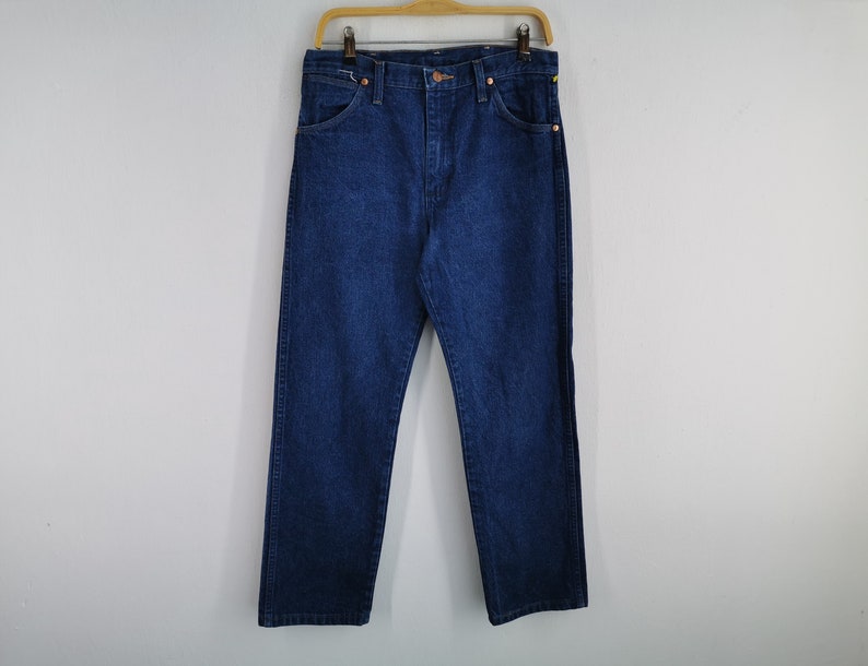 K&ouml;nnte beinhalten: Dunkelblaue Jeans aus Denim, die an einem Holzb&uuml;gel h&auml;ngen. Die Jeans haben ein klassisches F&uuml;nf-Taschen-Design mit geradem Bein. Der Denim scheint ein mittleres Gewicht zu haben, geeignet f&uuml;r den t&auml;glichen Gebrauch. Die Jeans werden vor einem schlichten wei&szlig;en Hintergrund pr&auml;sentiert.