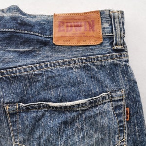 May include: Close-up of blue denim jeans with a brown leather patch. The patch features the brand name "EDWIN" in red lettering. The jeans have a back pocket and are size W32 L34.