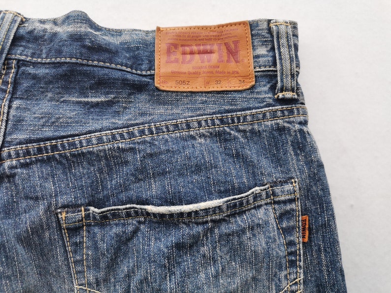 May include: Close-up of blue denim jeans with a brown leather patch. The patch features the brand name "EDWIN" in red lettering. The jeans have a back pocket and are size W32 L34.