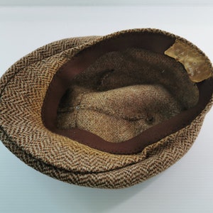 Op de afbeelding: Bruine tweed Kangol pet. De pet heeft een visgraatpatroon in bruin- en beigetinten. De binnenkant is gevoerd met bruine stof en heeft een bruine band langs de rand. Een Kangol-label is zichtbaar aan de binnenkant.