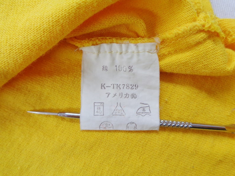 Op de afbeelding: Close-up van een geel kledinglabel met Japanse tekst en de tekst "100%" en "K-TK7829". Het label bevat ook verzorgingssymbolen en de tekst "アメリカ製". Een zilveren gereedschap zit in de afbeelding.