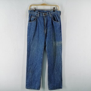 May include: Blue denim jeans hanging on a wooden hanger. The jeans have a classic five-pocket design and a straight leg cut. The denim has a faded appearance with some lighter areas, suggesting wear. The jeans are displayed against a plain white background.