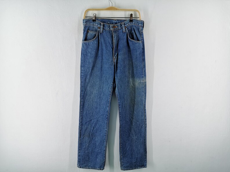 May include: Blue denim jeans hanging on a wooden hanger. The jeans have a classic five-pocket design and a straight leg cut. The denim has a faded appearance with some lighter areas, suggesting wear. The jeans are displayed against a plain white background.