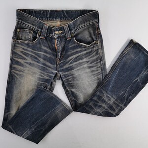 Op de afbeelding: Blauwe denim jeans met een vervaagde, versleten uitstraling. De jeans hebben een klassiek ontwerp met vijf zakken, een knoop- en ritssluiting en een licht uitlopende pijp. De stof vertoont slijtagepatronen.