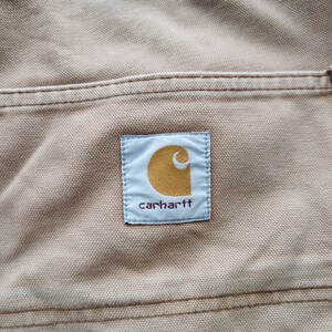 May include: Close-up of tan-colored fabric with a pocket and the Carhartt logo. The logo is a white rectangle with a gold-colored, stylized 'C' and the brand name in a dark font. The pocket has a reinforced edge with a metal rivet.