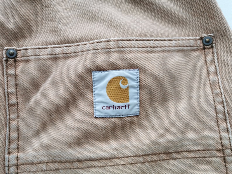 May include: Close-up of tan-colored fabric with a pocket and the Carhartt logo. The logo is a white rectangle with a gold-colored, stylized 'C' and the brand name in a dark font. The pocket has a reinforced edge with a metal rivet.