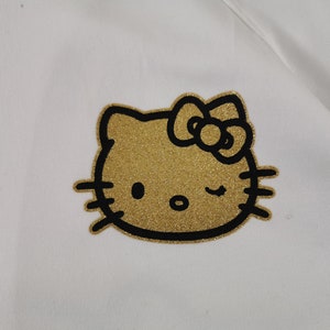 May include: A white fabric with a gold glitter Hello Kitty graphic. The Hello Kitty design is outlined in black and features a bow and winking eye. The fabric appears to be a soft material.