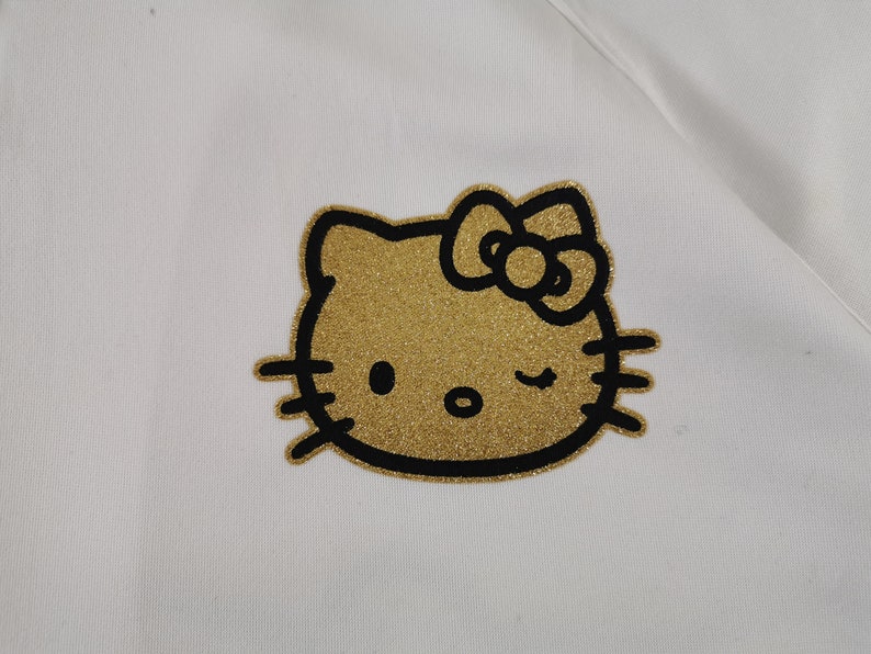 May include: A white fabric with a gold glitter Hello Kitty graphic. The Hello Kitty design is outlined in black and features a bow and winking eye. The fabric appears to be a soft material.