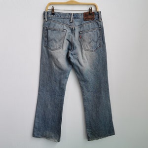 May include: Light blue denim jeans with a faded wash, hanging on a wooden hanger. The jeans have a classic five-pocket design, a leather patch, and a red tab on the back pocket. The jeans are displayed against a white background.