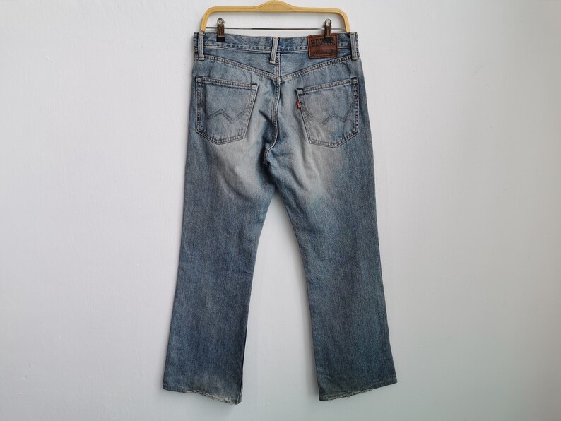 May include: Light blue denim jeans with a faded wash, hanging on a wooden hanger. The jeans have a classic five-pocket design, a leather patch, and a red tab on the back pocket. The jeans are displayed against a white background.