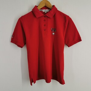 K&ouml;nnte beinhalten: Ein rotes Poloshirt mit kurzen &Auml;rmeln und Kragen. Das Shirt hat eine Knopfleiste mit drei Kn&ouml;pfen und ein kleines gesticktes Logo auf der Brust. Der &Auml;rmel hat ein kleines Logo. Das Shirt h&auml;ngt an einem Holzb&uuml;gel.
