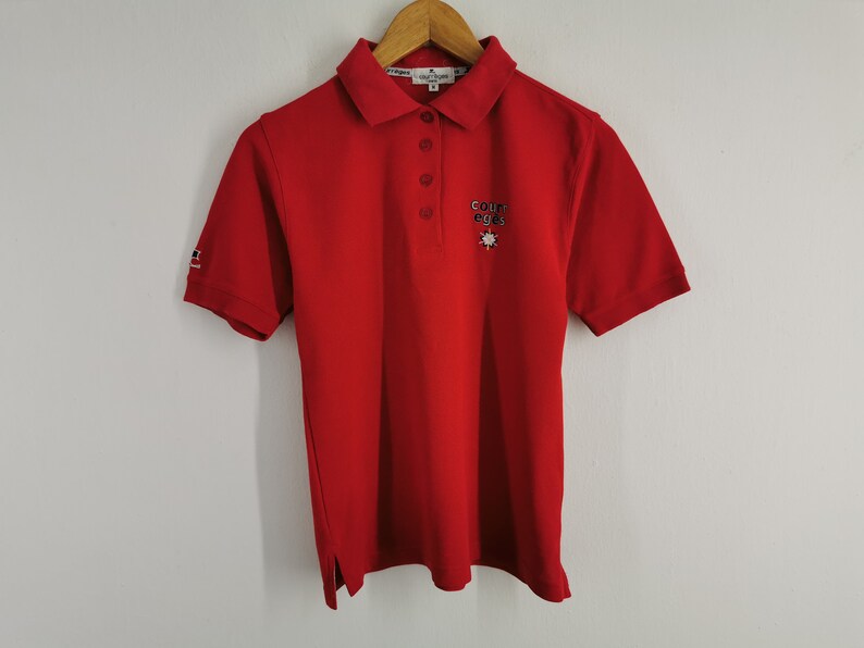 K&ouml;nnte beinhalten: Ein rotes Poloshirt mit kurzen &Auml;rmeln und Kragen. Das Shirt hat eine Knopfleiste mit drei Kn&ouml;pfen und ein kleines gesticktes Logo auf der Brust. Der &Auml;rmel hat ein kleines Logo. Das Shirt h&auml;ngt an einem Holzb&uuml;gel.