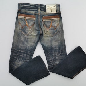 Pu&ograve; includere: Jeans in denim blu slavato con dettagli in pelle marrone sulle tasche posteriori. Presenta un design cucito decorativo sulle tasche posteriori e una toppa in pelle con un logo a teschio di toro.