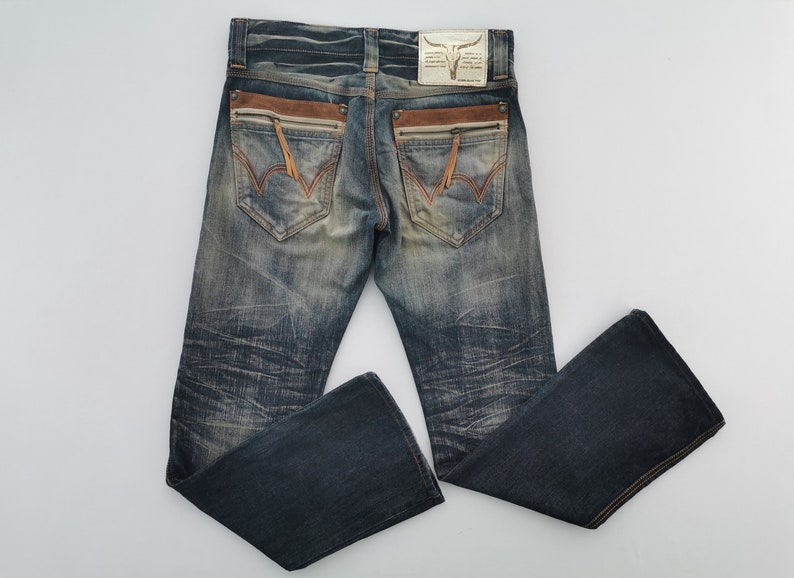 Pu&ograve; includere: Jeans in denim blu slavato con dettagli in pelle marrone sulle tasche posteriori. Presenta un design cucito decorativo sulle tasche posteriori e una toppa in pelle con un logo a teschio di toro.