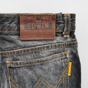 May include: Dark grey acid-wash denim jeans featuring a brown leather patch on the back waistband with the text EDWIN INTERNATIONAL BASIC. The jeans have orange contrast stitching, a back pocket, and a small yellow tag on the pocket edge.