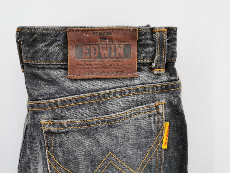 May include: Dark grey acid-wash denim jeans featuring a brown leather patch on the back waistband with the text EDWIN INTERNATIONAL BASIC. The jeans have orange contrast stitching, a back pocket, and a small yellow tag on the pocket edge.