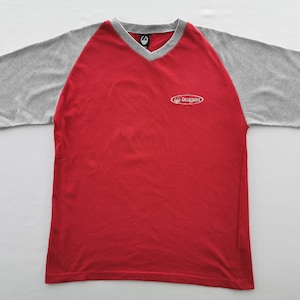 May include: Red and gray short-sleeve t-shirt with a V-neck. The shirt has a red body and gray sleeves. The word "Dragon" is printed in white on the chest. The shirt is made of a textured material.