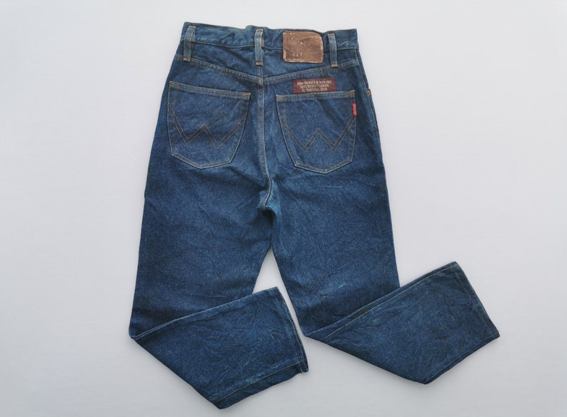 Puede incluir: Vaqueros de mezclilla azules con un dise&ntilde;o cl&aacute;sico de cinco bolsillos. Los vaqueros tienen un parche de cuero marr&oacute;n y una etiqueta roja en el bolsillo trasero. El logotipo de Wrangler es visible en los bolsillos traseros. Los vaqueros son de lavado medio.
