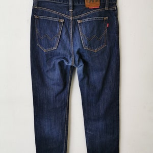 May include: Dark wash denim jeans with a classic five-pocket design. The jeans feature a straight leg cut and a leather patch with the brand name "EDWIN" on the back. The jeans are hanging on a metal hanger.