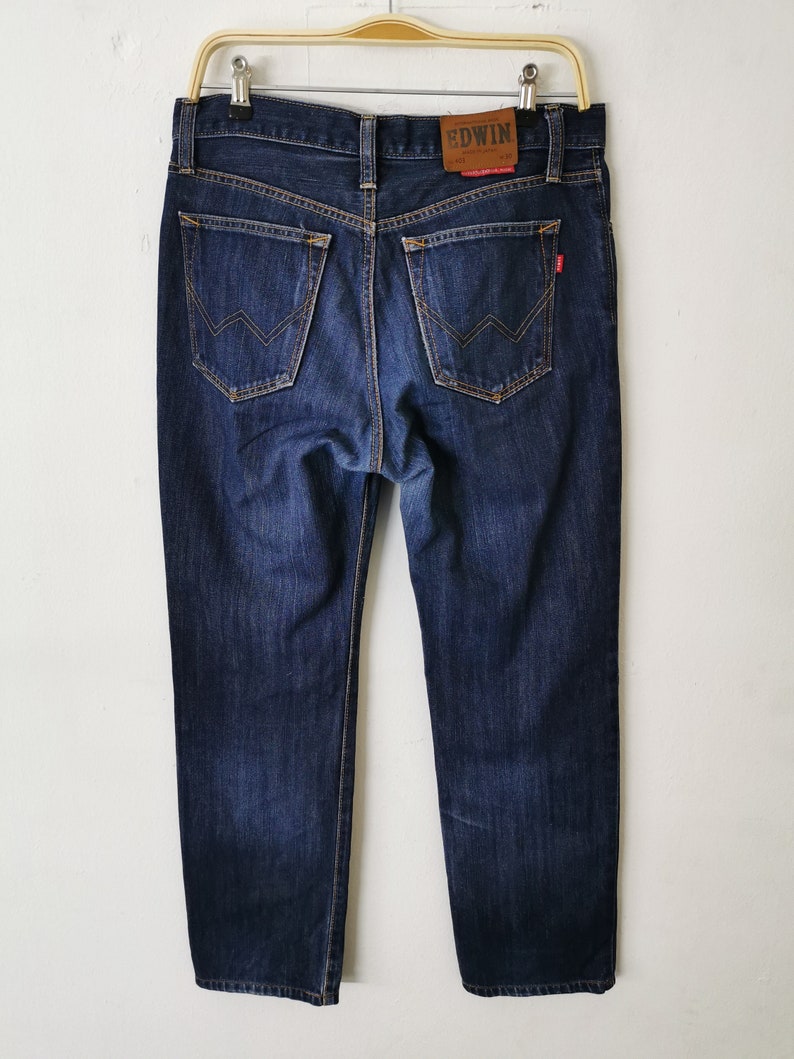 May include: Dark wash denim jeans with a classic five-pocket design. The jeans feature a straight leg cut and a leather patch with the brand name "EDWIN" on the back. The jeans are hanging on a metal hanger.