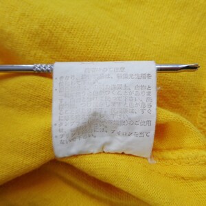 Op de afbeelding: Close-up van een felgele stof met een wit label eraan. Het label, vastgehouden door een zilveren speld, bevat Japanse tekst. De stof lijkt deel uit te maken van een kledingstuk.