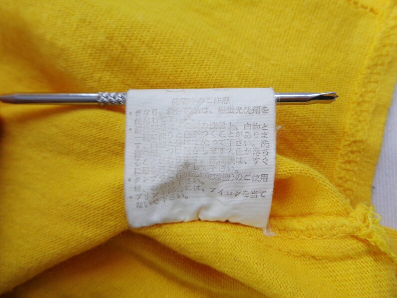 Op de afbeelding: Close-up van een felgele stof met een wit label eraan. Het label, vastgehouden door een zilveren speld, bevat Japanse tekst. De stof lijkt deel uit te maken van een kledingstuk.