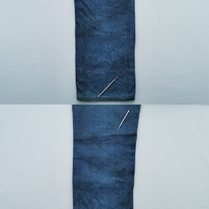 Puede incluir: Dos secciones de vaqueros de mezclilla azul se muestran verticalmente sobre un fondo gris claro. La mezclilla tiene un aspecto ligeramente descolorido con textura y pliegues sutiles. La imagen est&aacute; bien iluminada, destacando los detalles de la tela.