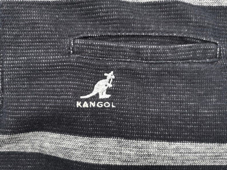 May include: Close-up of a black and gray striped fabric with a pocket. The white embroidered KANGOL logo featuring a kangaroo is visible. The fabric has a textured appearance.
