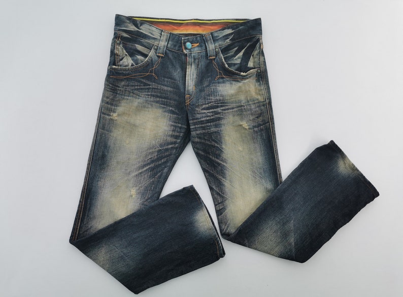 Pu&ograve; includere: Jeans di denim blu strappati con un lavaggio sbiadito e cuciture arancioni. I jeans hanno una chiusura con bottoni e un bottone turchese. I pantaloni in denim hanno un classico design a cinque tasche e una vestibilit&agrave; rilassata.