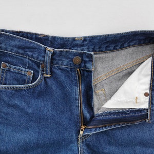 Puede incluir: Primer plano de unos vaqueros de mezclilla azules con una cremallera parcialmente desabrochada. Los vaqueros tienen un dise&ntilde;o cl&aacute;sico de cinco bolsillos, con un peque&ntilde;o bolsillo para monedas y remaches. El forro interior es visible.