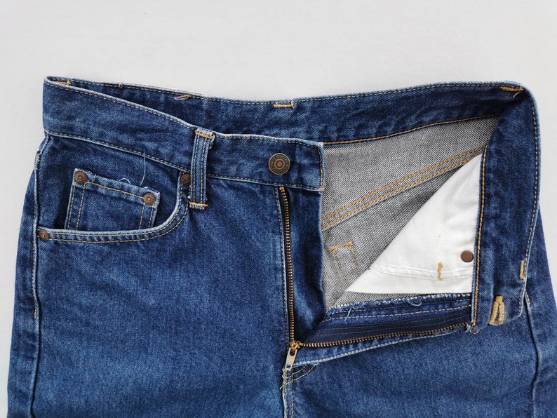 Puede incluir: Primer plano de unos vaqueros de mezclilla azules con una cremallera parcialmente desabrochada. Los vaqueros tienen un dise&ntilde;o cl&aacute;sico de cinco bolsillos, con un peque&ntilde;o bolsillo para monedas y remaches. El forro interior es visible.