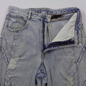 May include: Light blue acid wash denim jeans with a partially unzipped fly. The jeans have a distressed look with ripped areas and unique seam detailing. The pockets are visible, and the overall style is vintage.