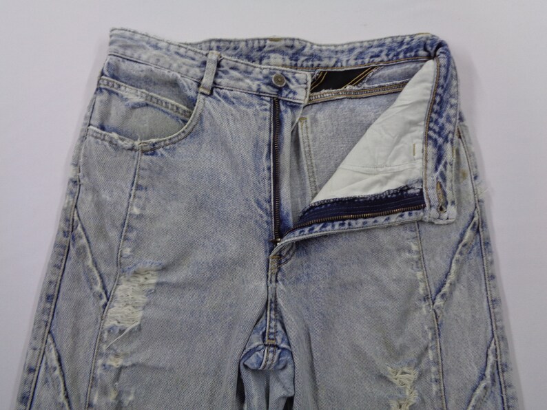 May include: Light blue acid wash denim jeans with a partially unzipped fly. The jeans have a distressed look with ripped areas and unique seam detailing. The pockets are visible, and the overall style is vintage.