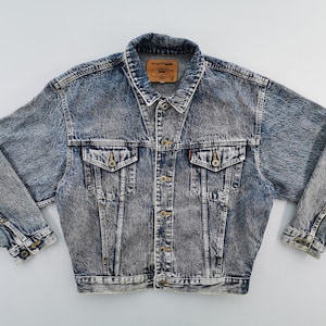 May include: A vintage acid wash denim jacket. It features a button-up front, two chest pockets, and two side pockets. The jacket has a classic collar and buttoned cuffs. The denim is a faded blue with a mottled, textured appearance. A brown label is visible on the back of the collar.