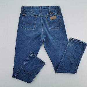 May include: A pair of medium-wash blue denim jeans featuring a classic five-pocket design, a leather-like patch on the back right pocket, and visible dark stains on the lower legs. The jeans include belt loops and copper-colored rivets.