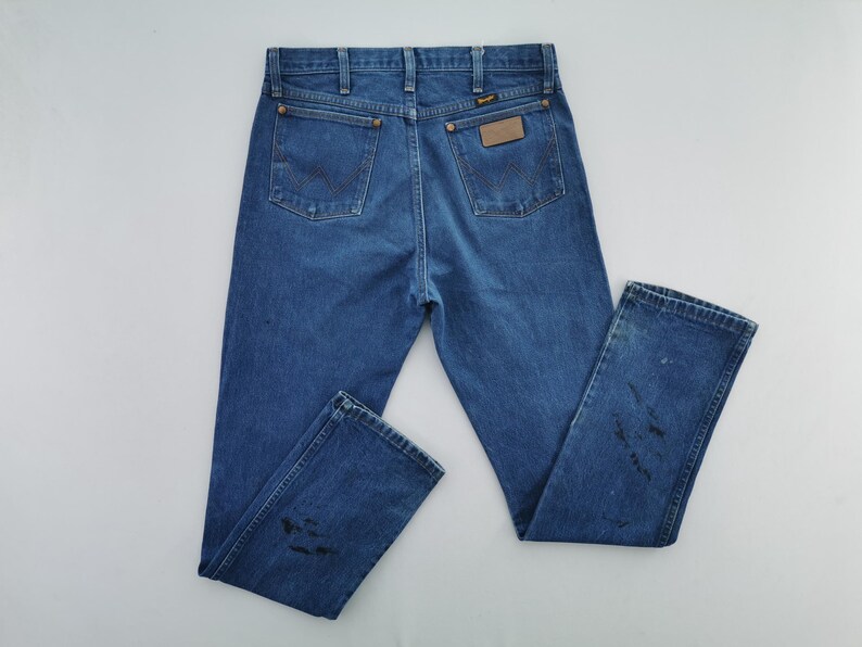 May include: A pair of medium-wash blue denim jeans featuring a classic five-pocket design, a leather-like patch on the back right pocket, and visible dark stains on the lower legs. The jeans include belt loops and copper-colored rivets.