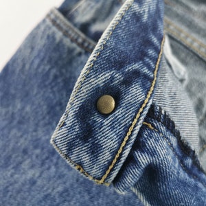 Puede incluir: Primer plano de unos vaqueros azules, mostrando el bot&oacute;n y los detalles de las costuras. Los vaqueros tienen un dise&ntilde;o cl&aacute;sico con cierre de bot&oacute;n y costuras amarillas visibles. La tela tiene un aspecto texturizado.