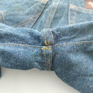 Puede incluir: Primer plano de unos vaqueros de mezclilla azules, que muestran la cintura y los detalles de las costuras. Los vaqueros tienen un dise&ntilde;o cl&aacute;sico con costuras visibles y una tela texturizada. Las costuras son de color amarillo contrastante.