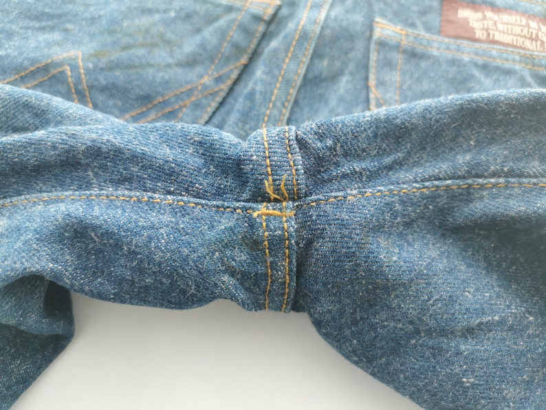 Puede incluir: Primer plano de unos vaqueros de mezclilla azules, que muestran la cintura y los detalles de las costuras. Los vaqueros tienen un dise&ntilde;o cl&aacute;sico con costuras visibles y una tela texturizada. Las costuras son de color amarillo contrastante.